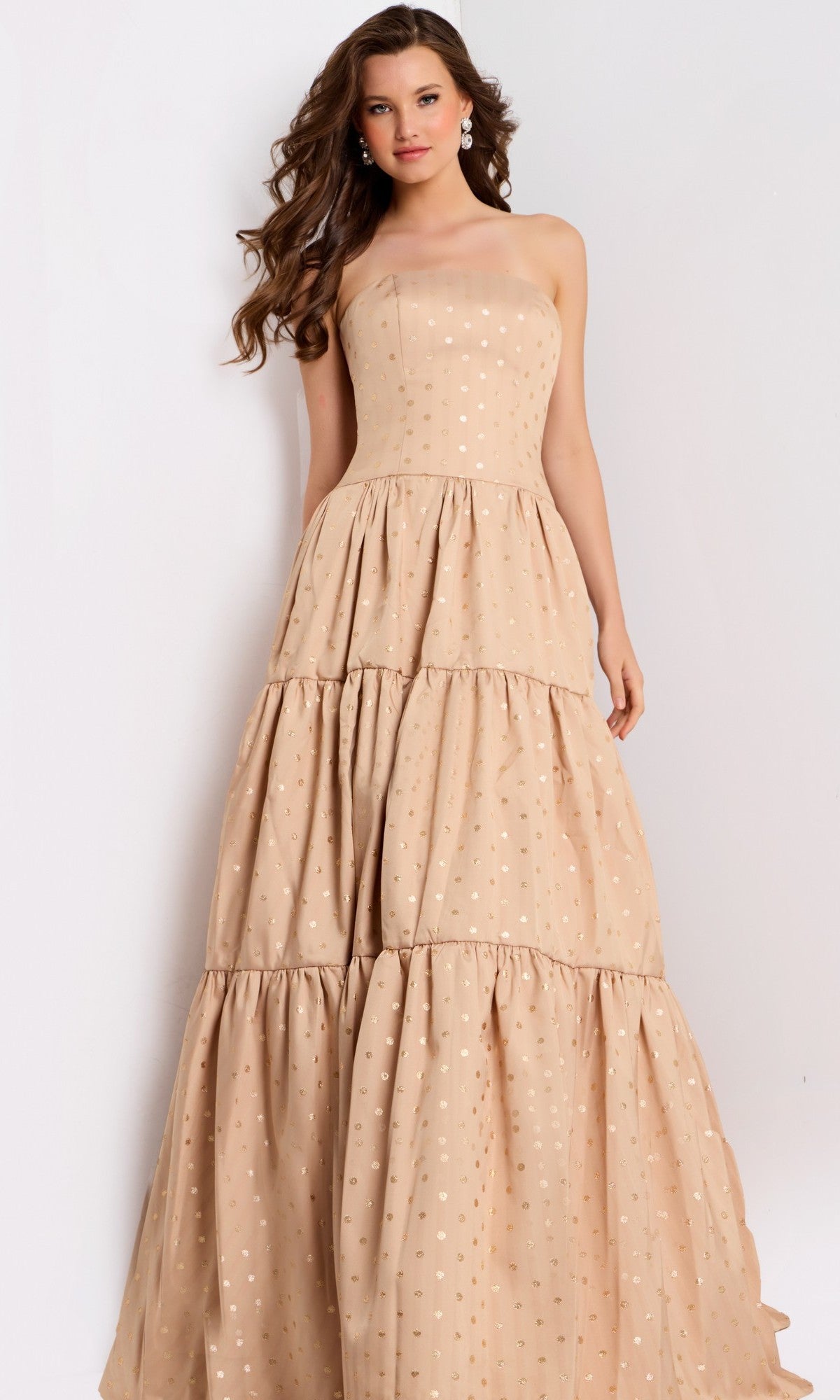A woman stands against a plain background wearing the Jovani 49341 long prom dress in gold, featuring a strapless design with subtle polka dots and tiered layers.